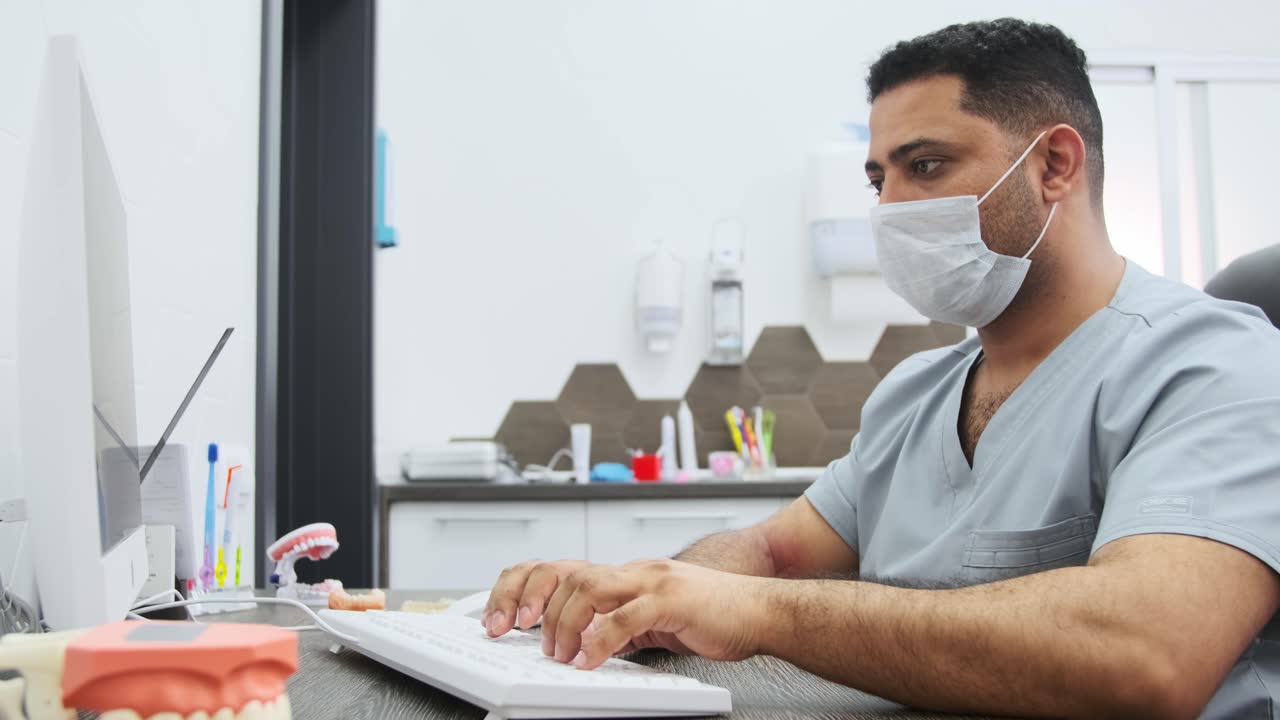 A dentist in scrubs types at a computer in the practice.
